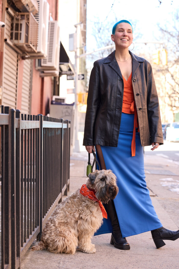 A smiling person with short blue hair walking a small fluffy dog on a city sidewalk